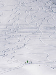 zaśnieżony stok, na którym widać ślady pozostawione przez narciarzy, u dołu stoku małe postaci dwojga ludzi, jeden z nich podnosi ręce to góry w zachwycie