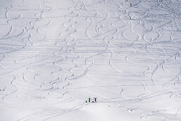 zaśnieżony stok, na którym widać ślady pozostawione przez narciarzy, u dołu stoku małe postaci dwojga ludzi, jeden z nich podnosi ręce to góry w zachwycie